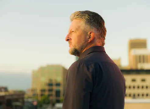 Side profile of a man with gray hair and beard looking into the distance over a cityscape.