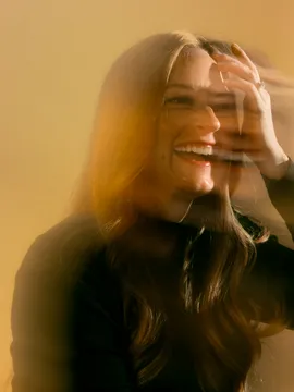 Smiling woman with long hair in black sweater touching her face against a yellow background.