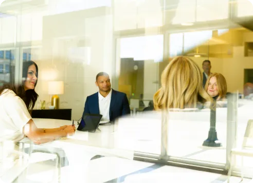 Four professionals in a bright office meeting around a large table with laptops and smiles.