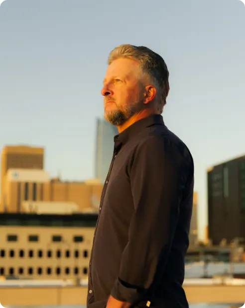 Man with beard and short hair in dark shirt standing and looking into distance on city rooftop at sunset.