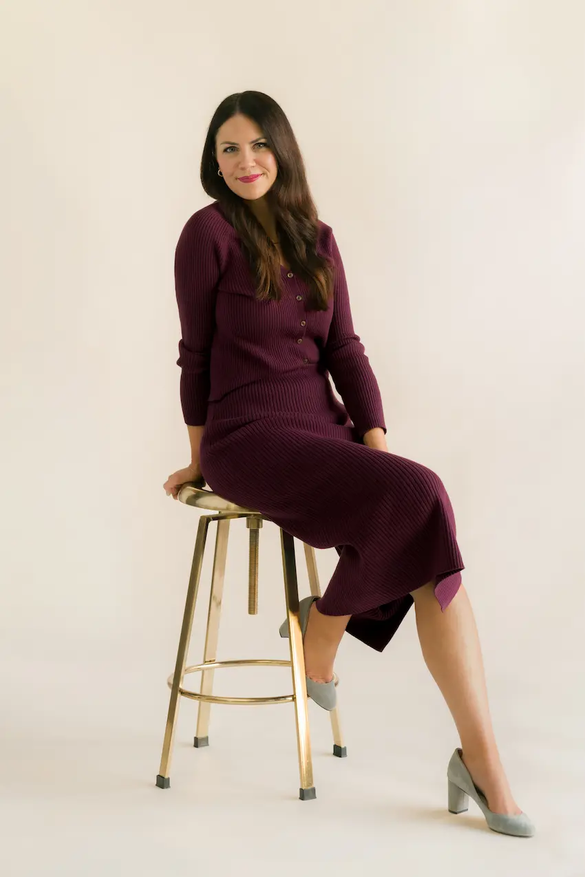 Woman with long dark hair in a purple ribbed dress and gray heels perched on a brass stool.