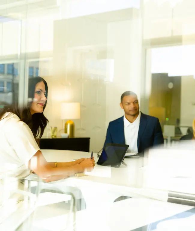 Two professionals sitting at a white table in a modern office meeting room with a tablet device.