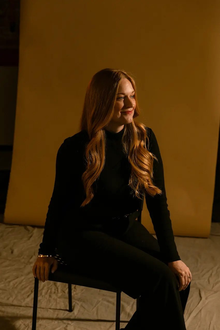 Woman with long wavy hair sitting on a chair against a mustard yellow backdrop, wearing black.