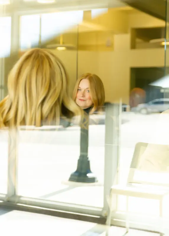 Reflection of a woman with blonde hair sitting in a bright room with glass windows.