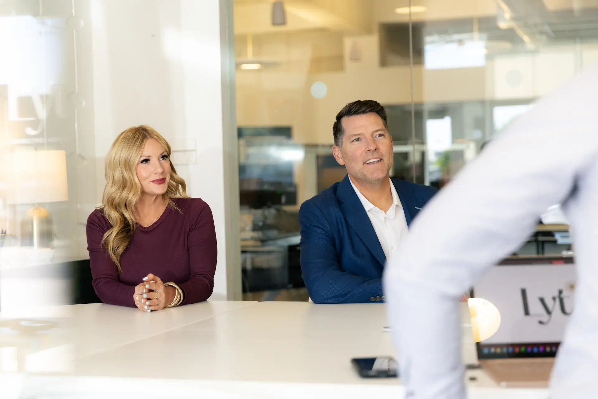 Man in blue jacket and woman in purple shirt sitting at a table in a bright office setting.