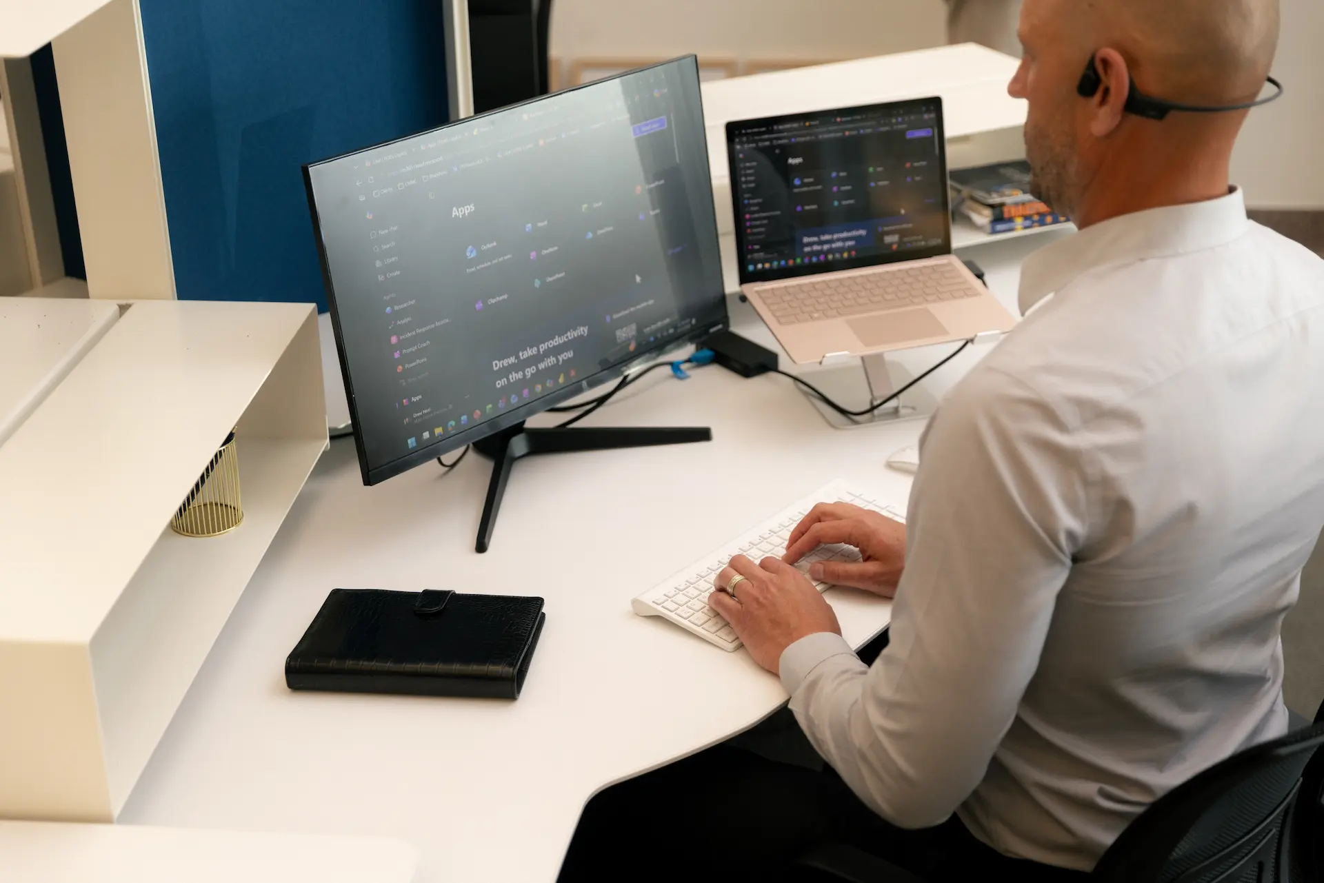 Man in headset typing on keyboard with curved monitor and laptop displaying productivity apps.