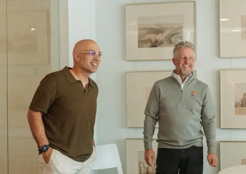 Two men smiling and standing in a room with framed artwork on the wall behind them.