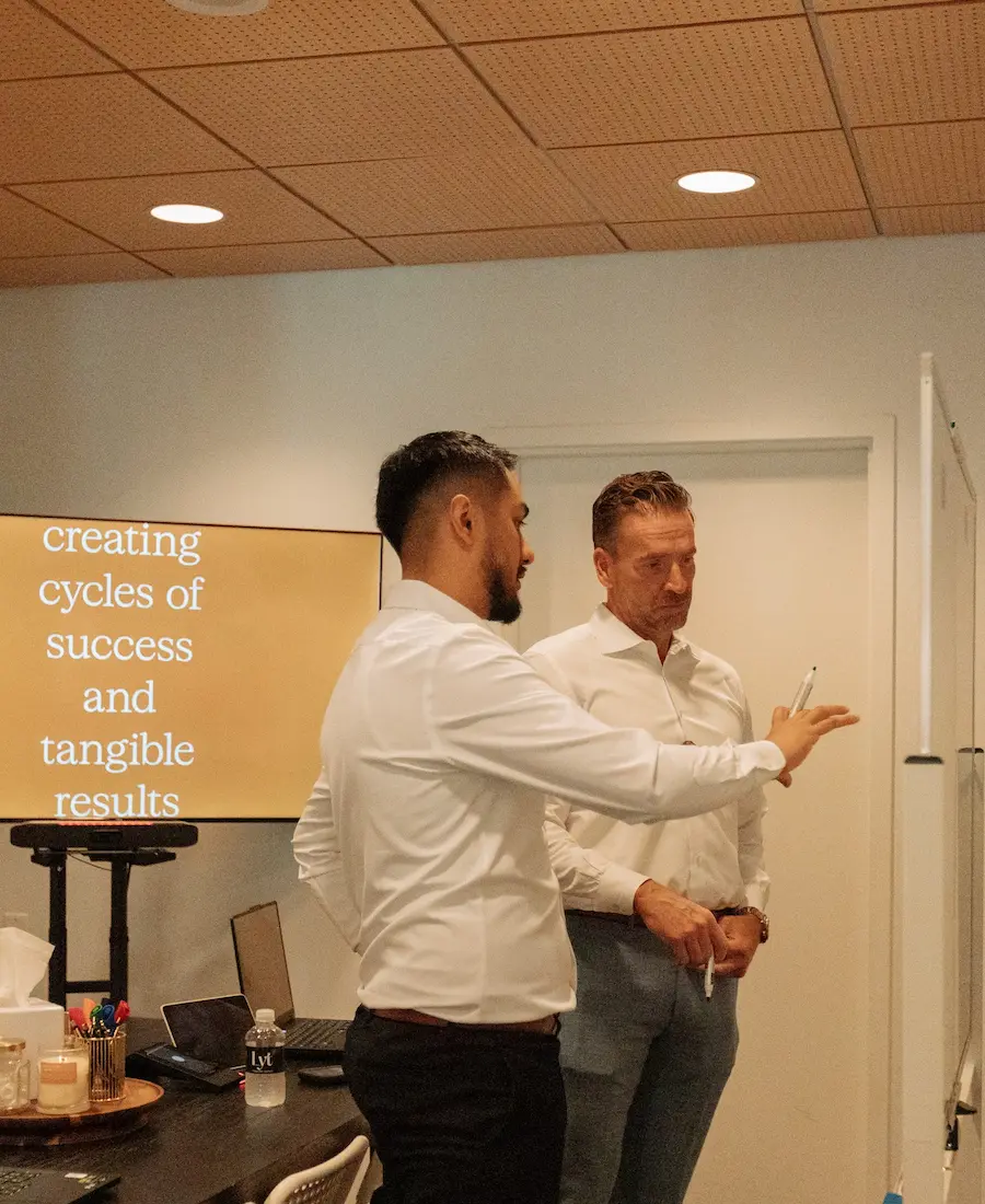 Two men in white shirts discussing and pointing at a whiteboard in a modern office setting.