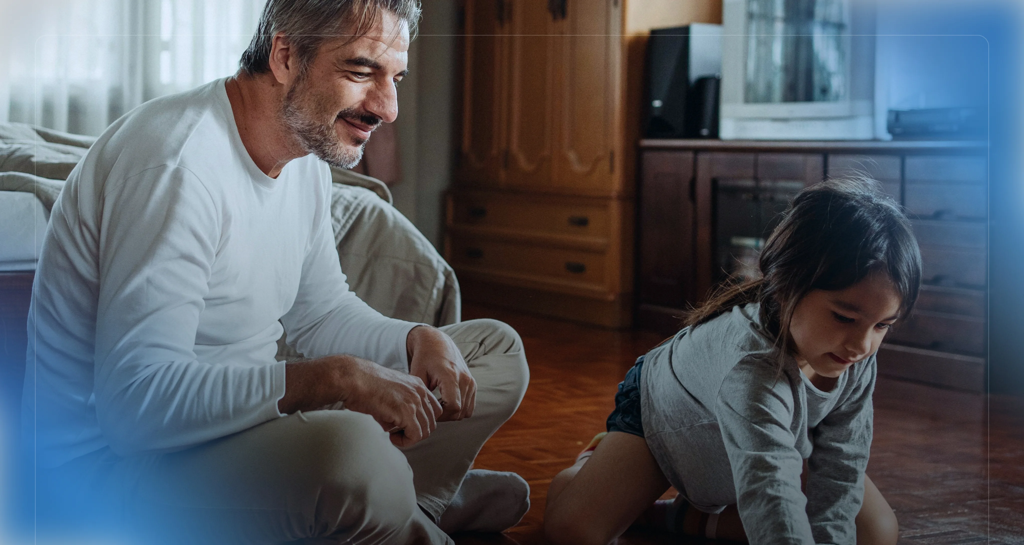 Un homme souriant assis par terre regarde une petite fille qui joue à côté de lui à l'intérieur.