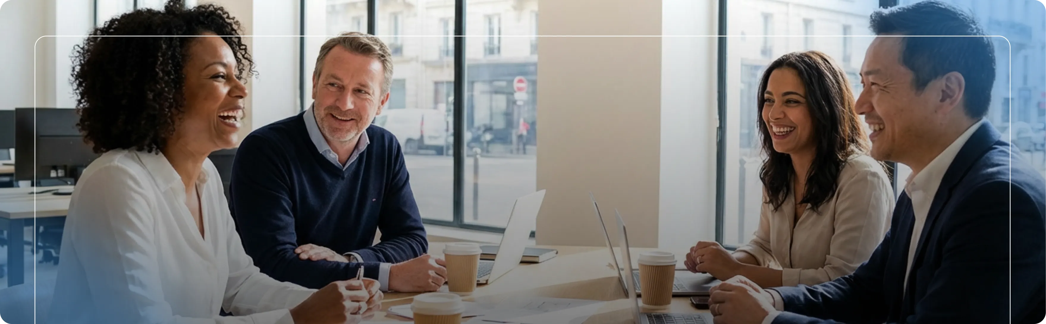 Quatre professionnels souriants discutant autour d'une table dans un bureau lumineux avec des ordinateurs portables.
