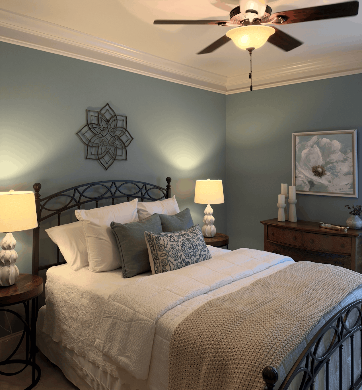 Cozy bedroom with a metal-framed bed dressed in white and beige bedding, accented with green and patterned pillows, flanked by two white lamps on wooden side tables against light blue walls with decorative metal wall art and a floral painting above a wooden dresser.