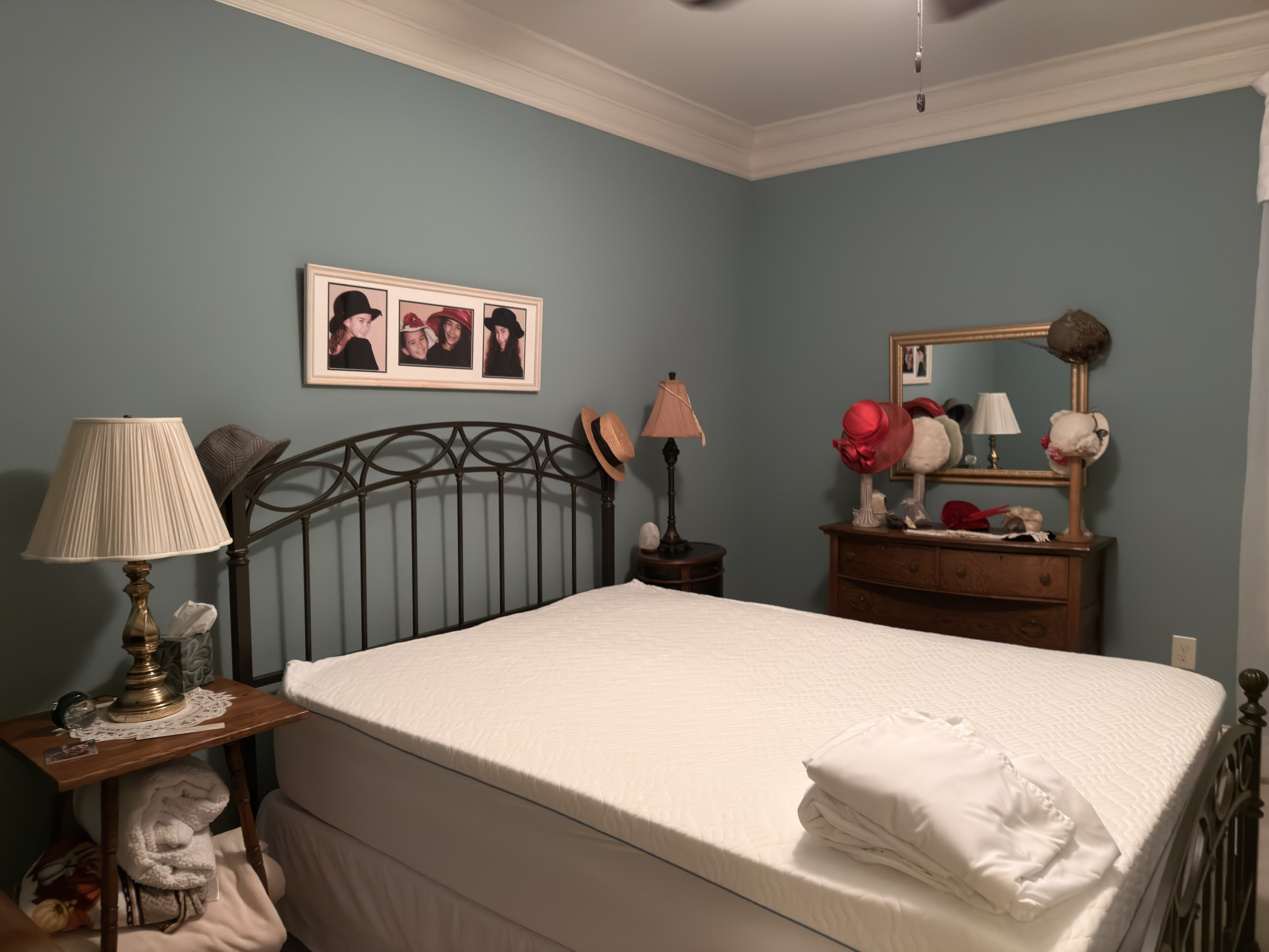 Bedroom with greenish-gray walls featuring a metal bed frame with white bedding and a folded sheet on top, a wooden nightstand with a brass lamp and tissues, and a wooden dresser with a mirror and multiple hats displayed on stands.