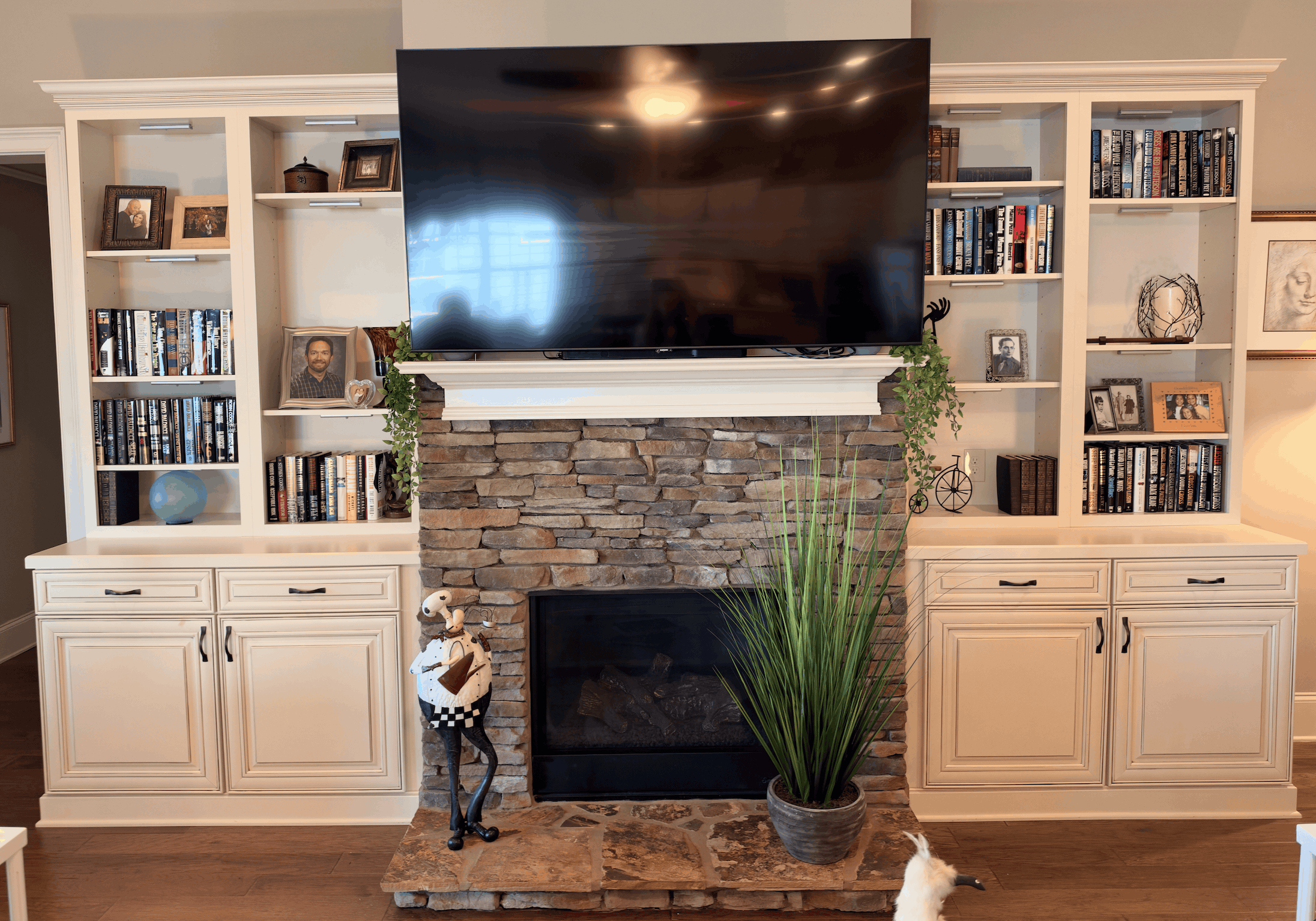 Living room fireplace with stacked stone surround and a mounted flat-screen TV above; white built-in shelves and cabinets flank the fireplace, holding books, framed photos, and decorative items; a tall green plant in a pot and a chef figurine stand on the hearth.