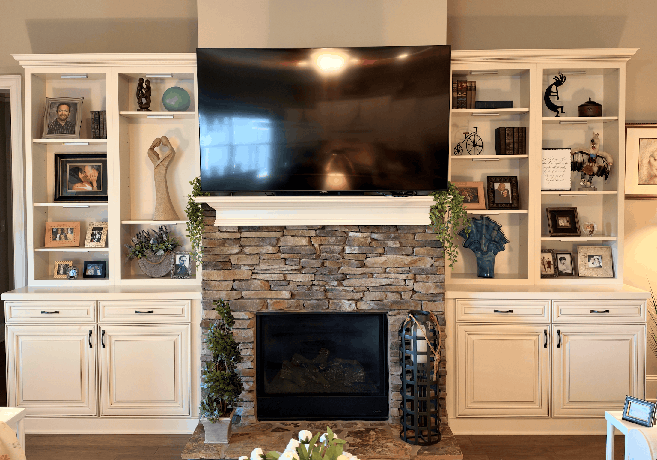 Stone fireplace with a large flat-screen TV mounted above it, flanked on both sides by white built-in shelves and cabinets decorated with framed photos, sculptures, books, plants, and decorative objects.