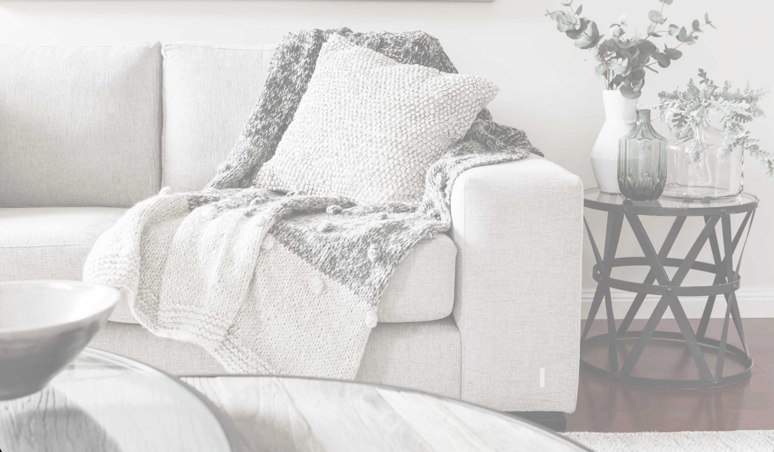 Light beige sofa with a knitted textured pillow and a gray and white knitted throw blanket draped over the armrest, next to a round glass-top black metal side table holding three vases with green leafy branches.