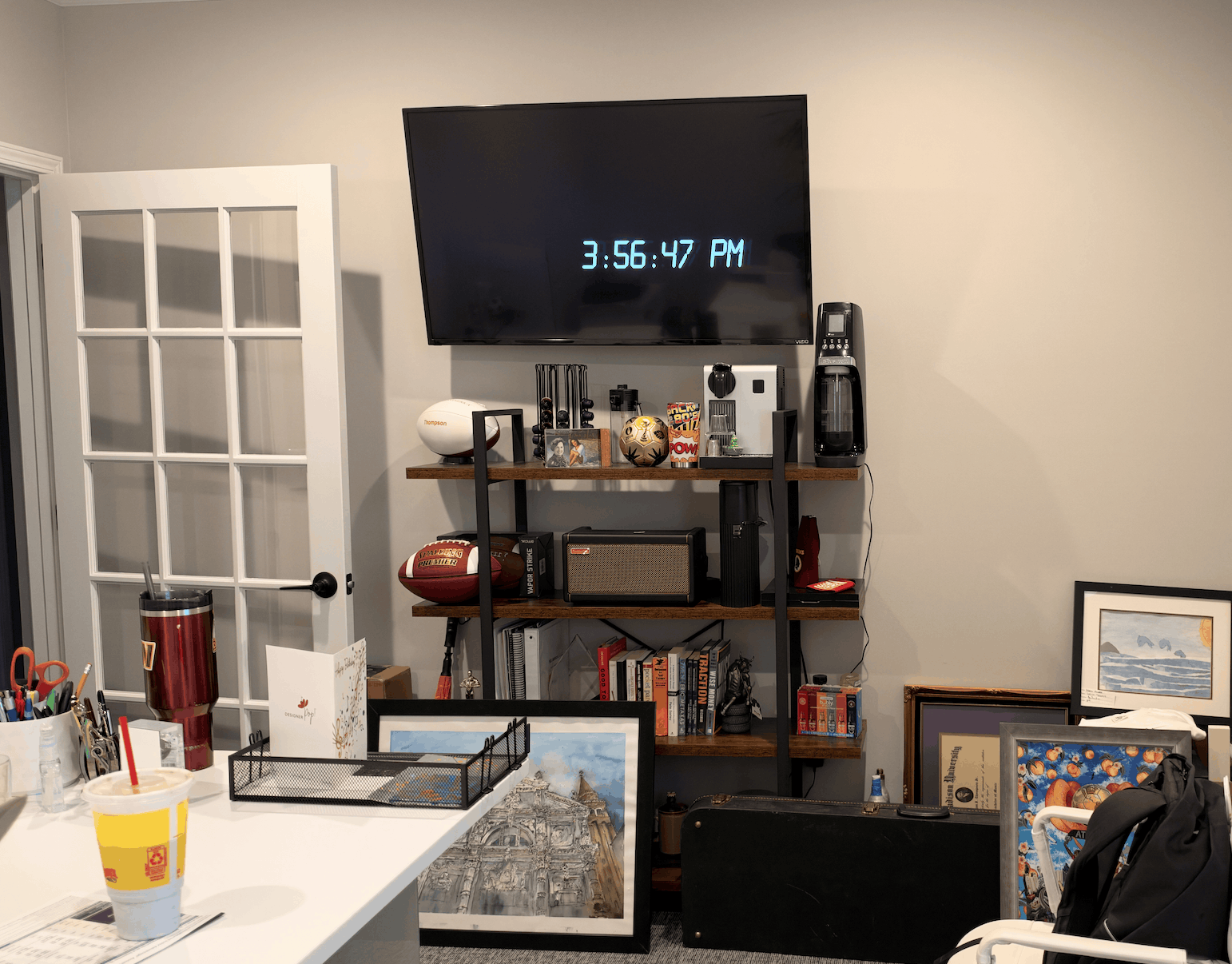 Office interior with a white desk holding pens, scissors, a yellow drink cup with a red straw, and a mesh tray. Behind the desk is a black metal and wood shelving unit against a beige wall, featuring a mounted flat-screen TV showing a digital clock reading 3:56:47 PM. The shelves display sports memorabilia including two American footballs, framed photos, books, a speaker, a coffee machine, and various items. The floor in front of the shelves has framed artwork and a black case. A white chair with a black jacket is in the right foreground, and a white door with glass panels is open on the left.