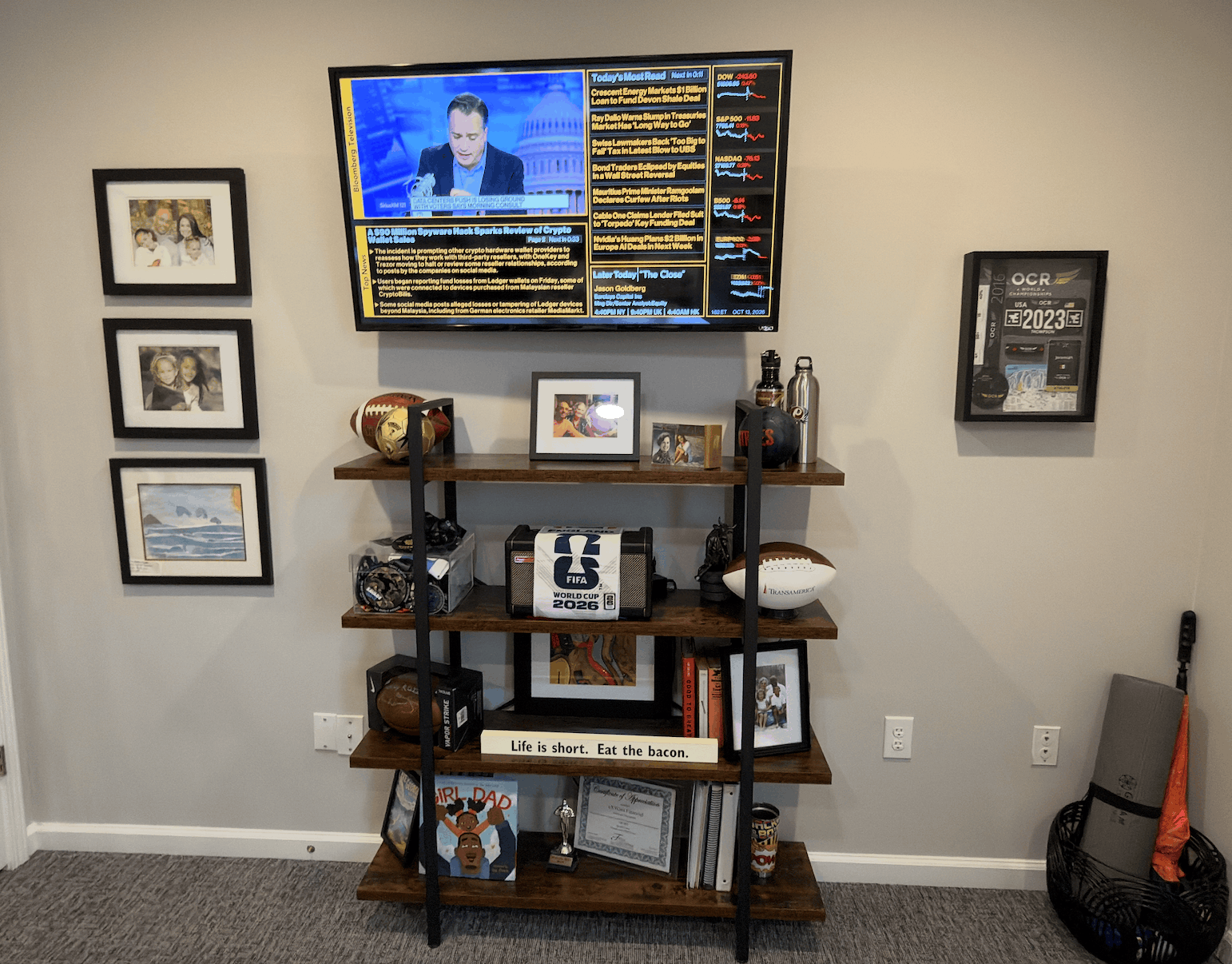 Office wall has a mounted TV displaying financial news and stock market updates. Below is a wooden shelf with four levels holding framed photos, footballs, sports memorabilia, books, a FIFA 2026 item, and a sign that reads 'Life is short. Eat the bacon.' To the left are three framed pictures vertically aligned on the wall. To the right, a framed OCR certificate and a basket on the floor containing an exercise mat and an orange umbrella.