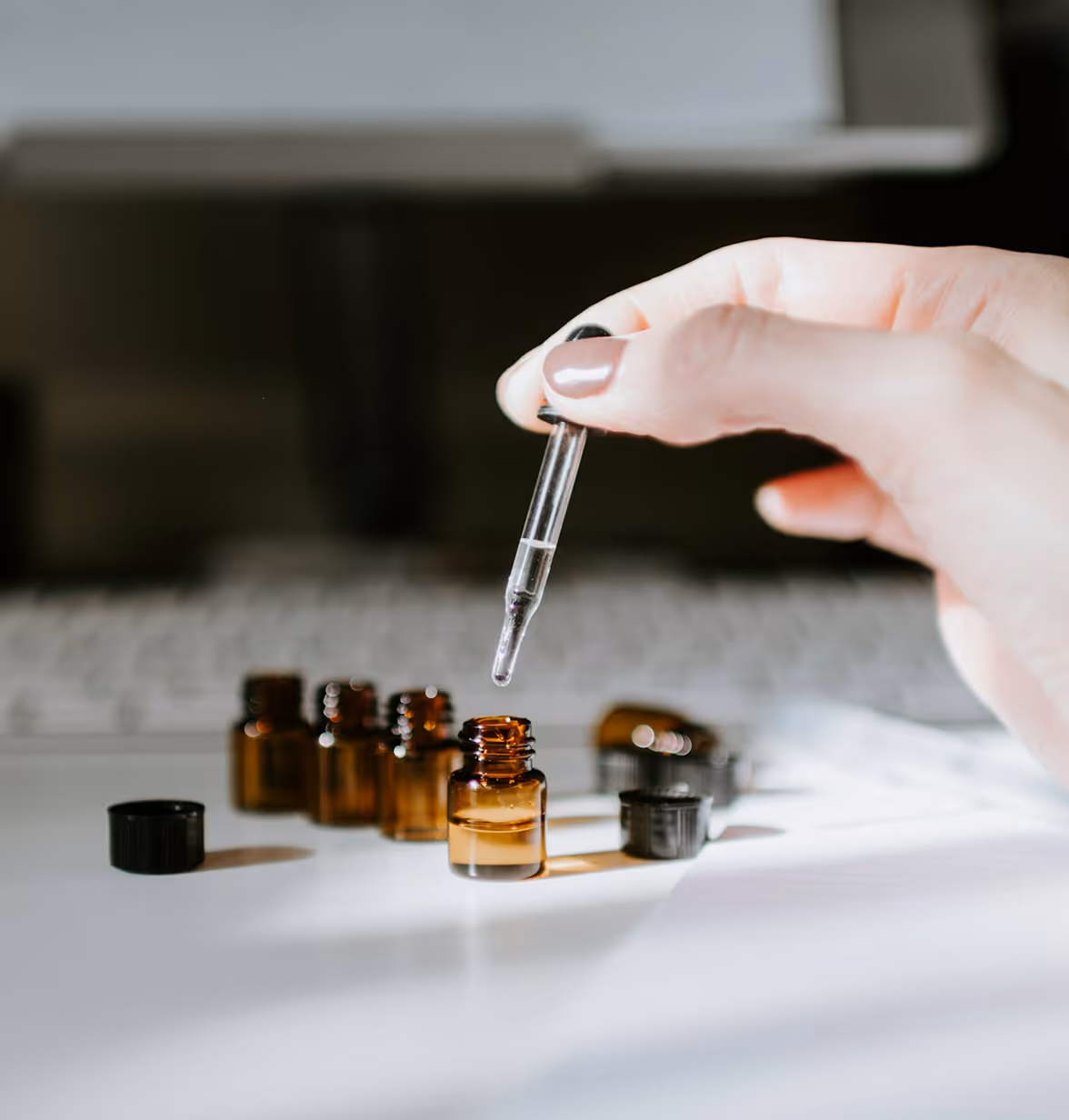 Hand holding a dropper above a small amber glass bottle with other bottles and caps nearby.