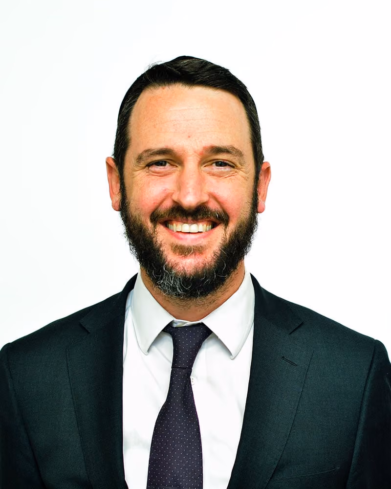 Smiling man with short dark hair and beard wearing a dark suit, white shirt, and patterned tie.