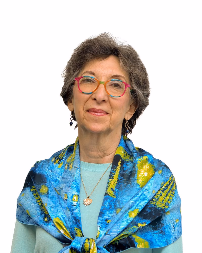 Older woman wearing multicolored glasses and a blue patterned scarf over a light shirt, smiling gently.