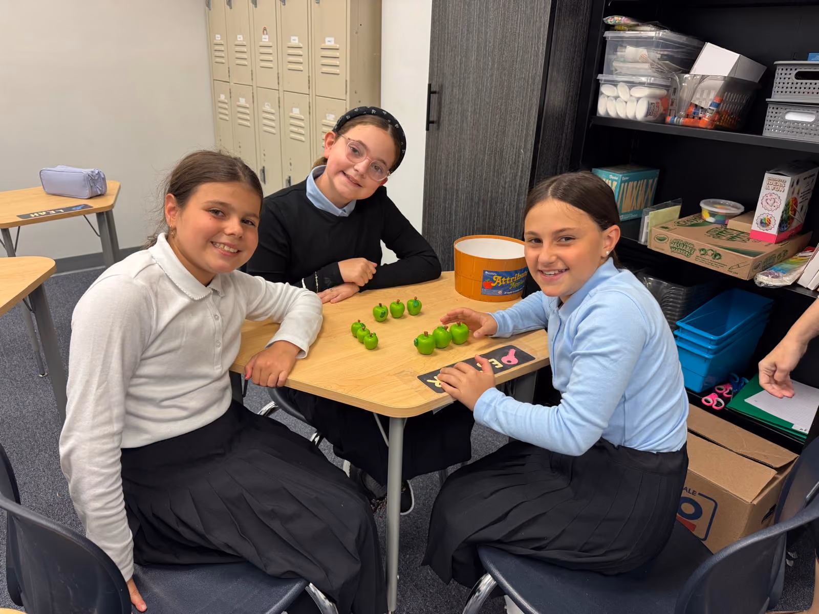Three girls in school uniforms seated around a table with small green apple toys arranged in rows.