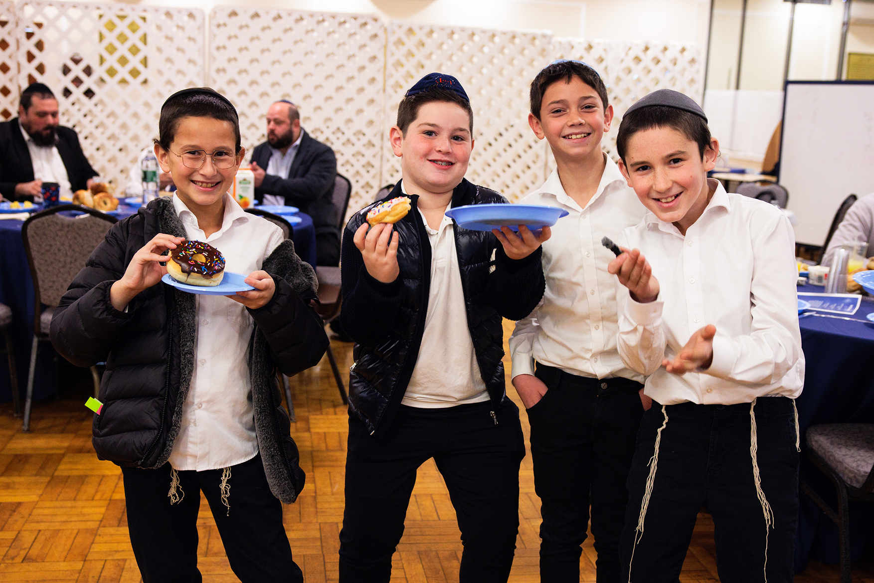 Four smiling boys wearing yarmulkes holding donuts and blue plates at a gathering.