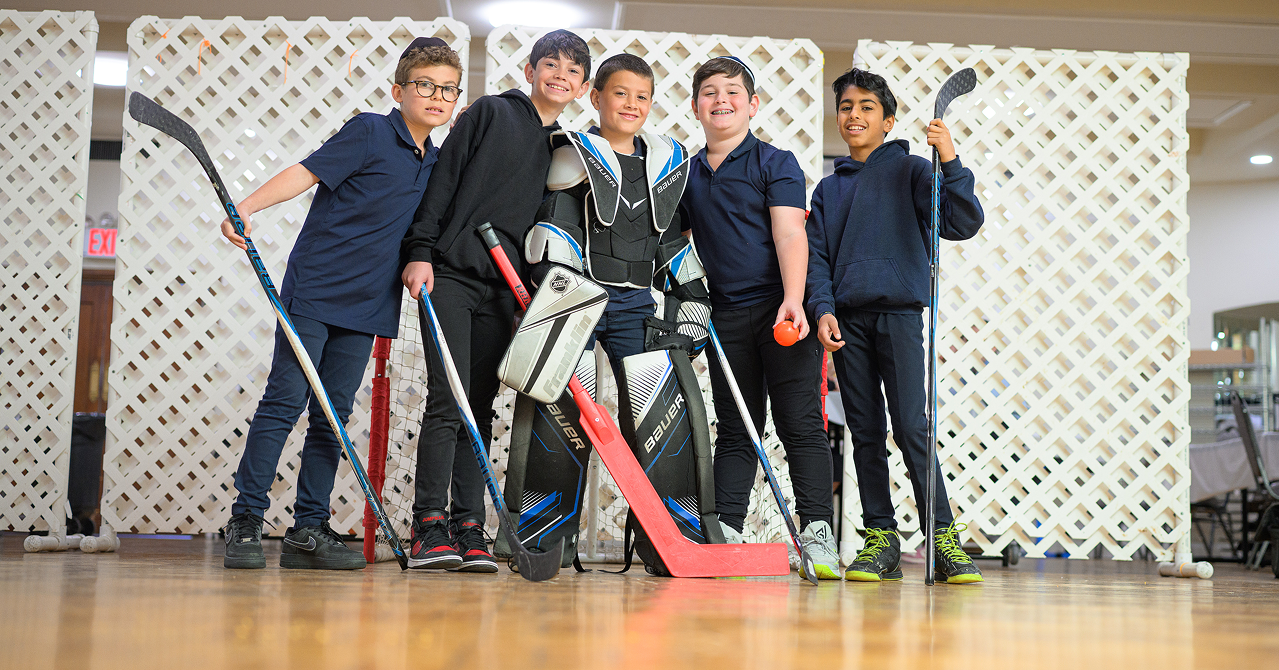 Five boys smiling with hockey sticks, one in goalie gear, standing indoors on wood floor.