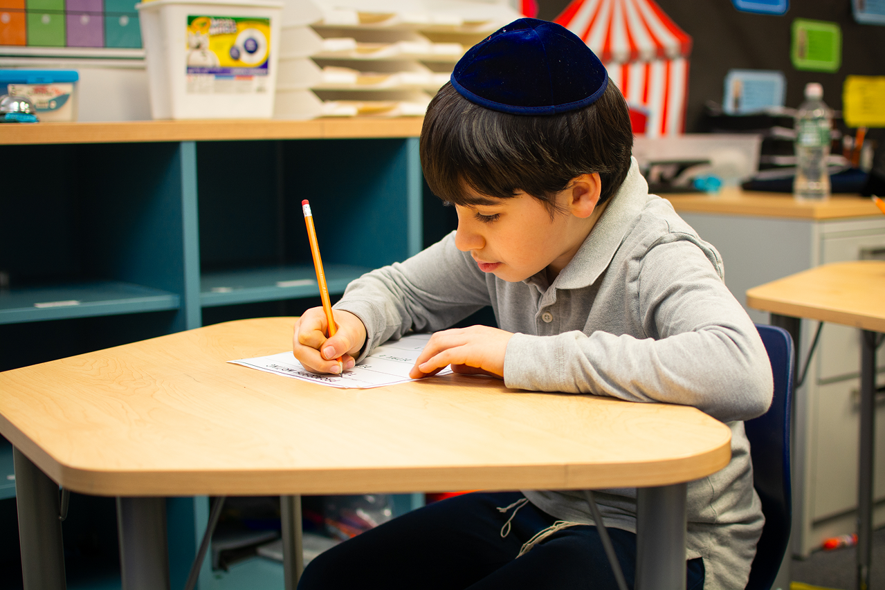 Young boy wearing a kippah writing with a pencil on paper at a school desk.