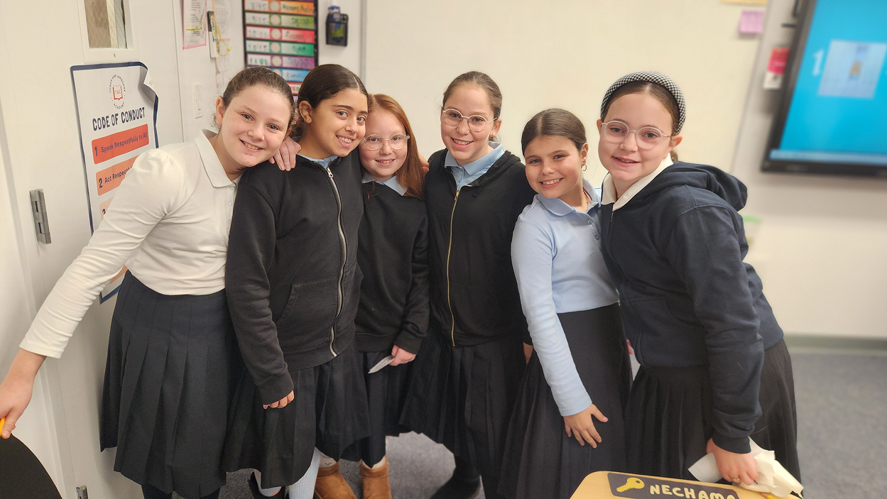Six smiling girls standing close indoors, wearing school uniforms with skirts and sweatshirts.