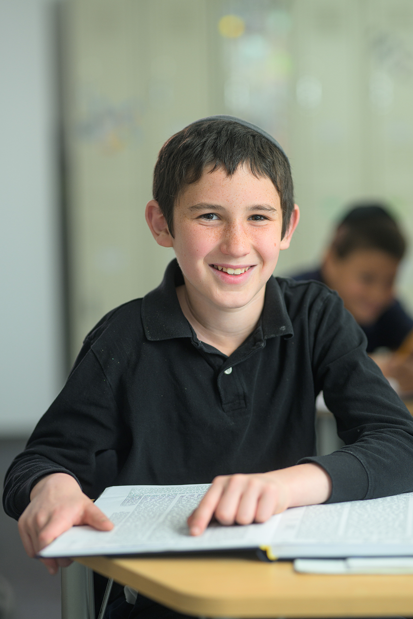 Smiling boy wearing a black shirt and kippah reading a book at a desk in a classroom.