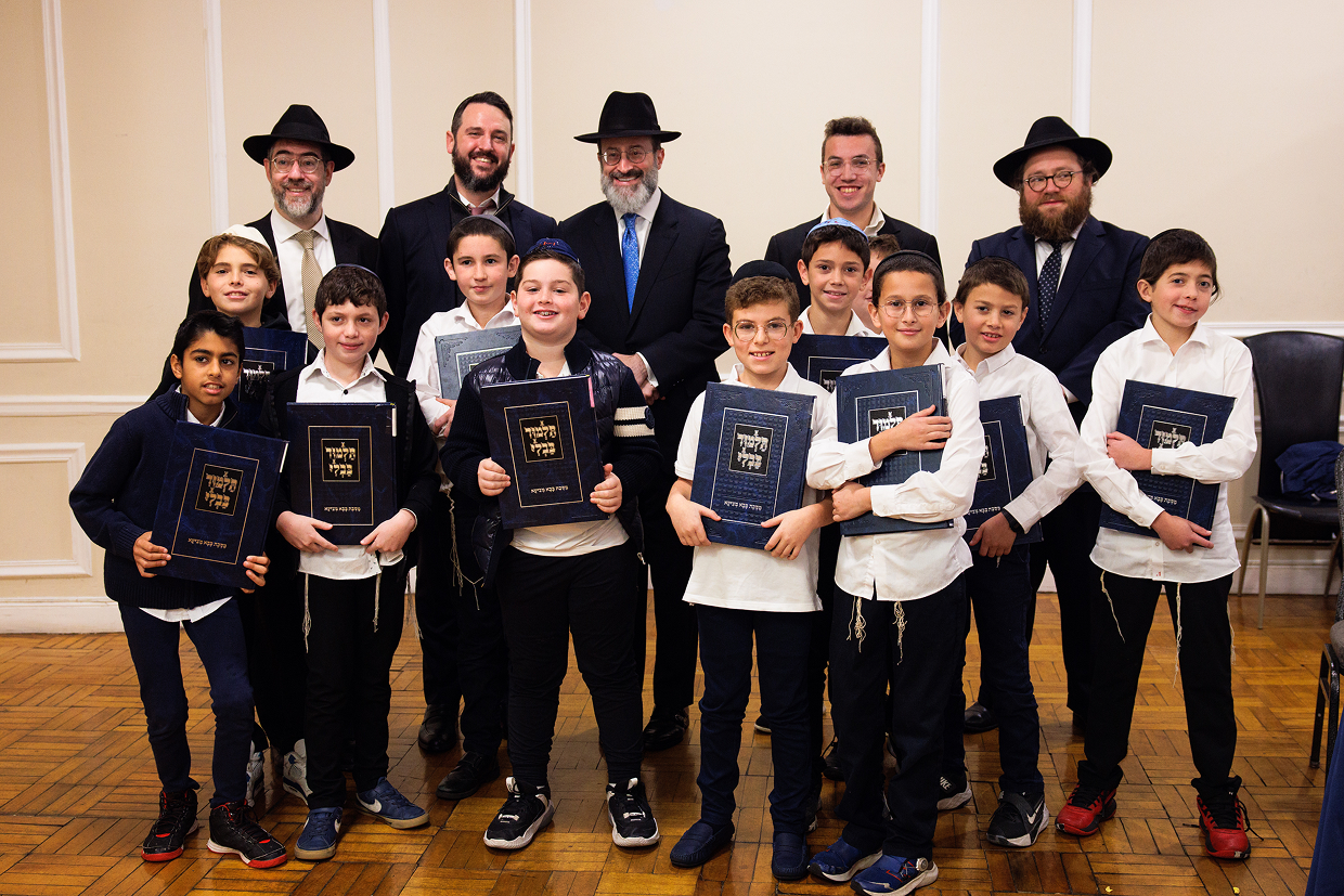 Group of boys and men in formal attire holding blue books posing indoors on a wooden floor.