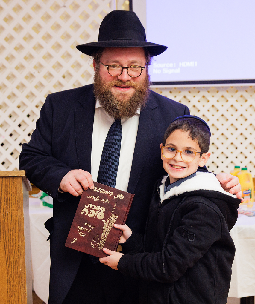 Man in black suit and hat and boy in glasses holding a book with Hebrew text smiling indoors.