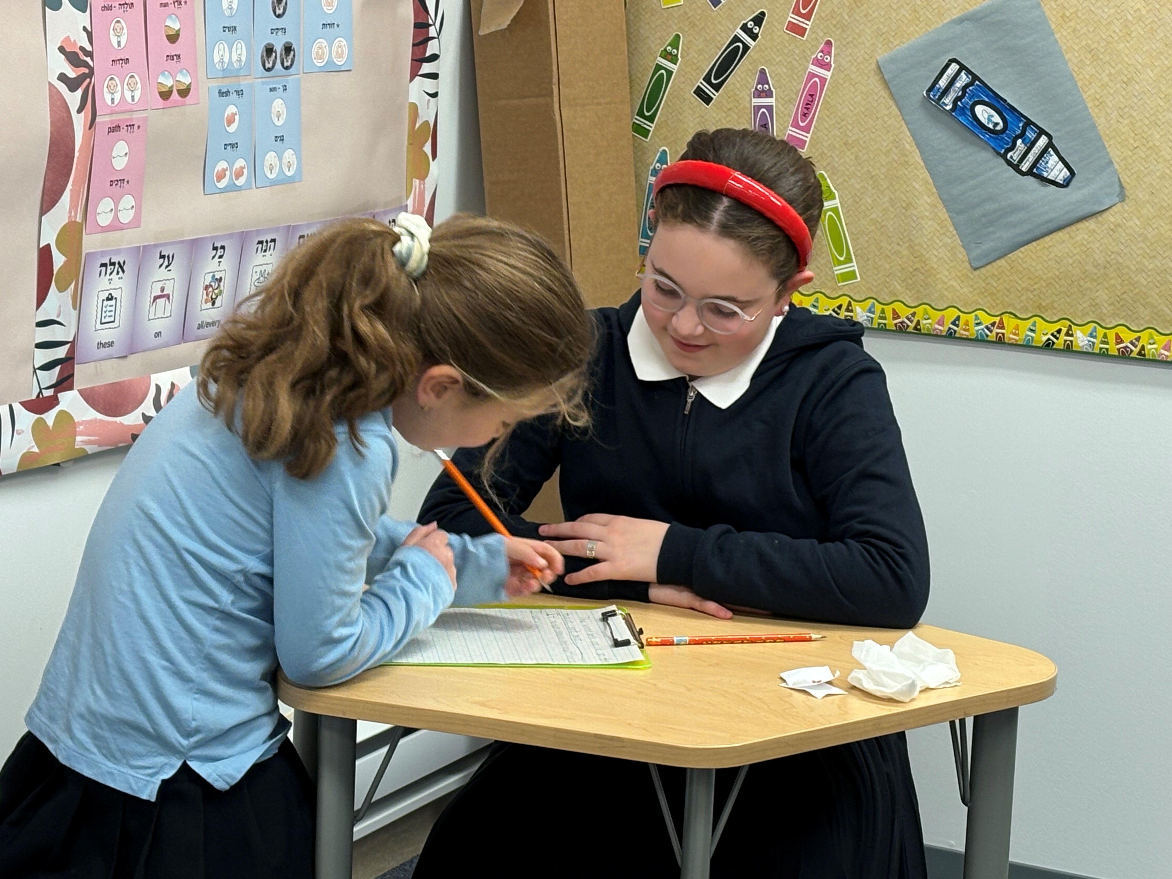Two girls working together at a classroom table with pencils and writing on a clipboard.