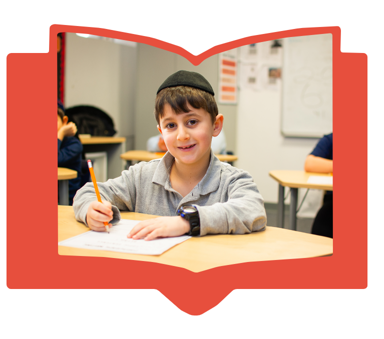 Young boy wearing a kippah writing with pencil at desk in classroom with smile.