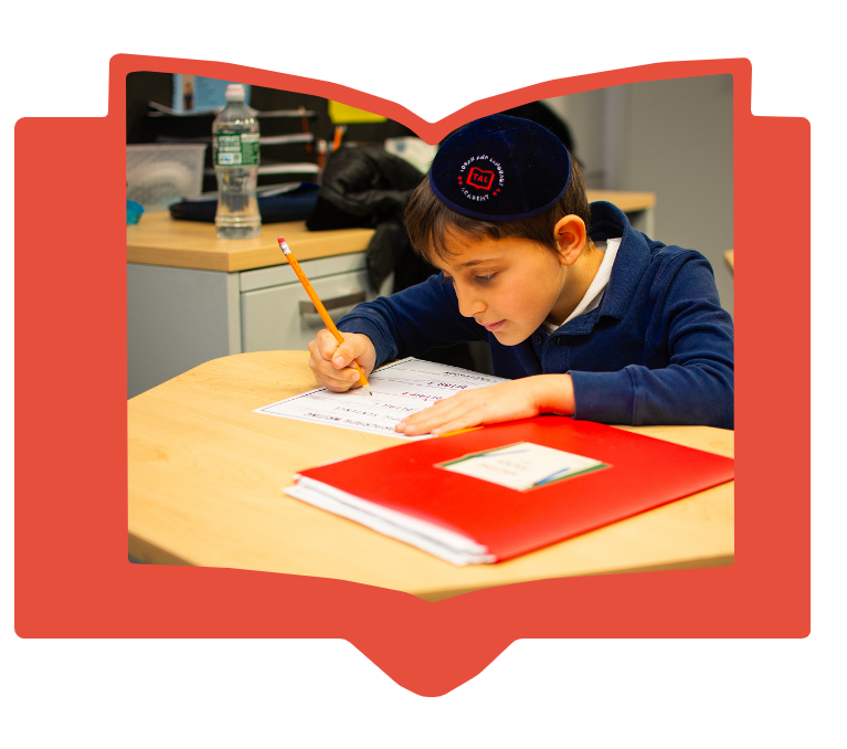 Young boy wearing a kippah writing with pencil on paper at a desk with a red folder nearby.