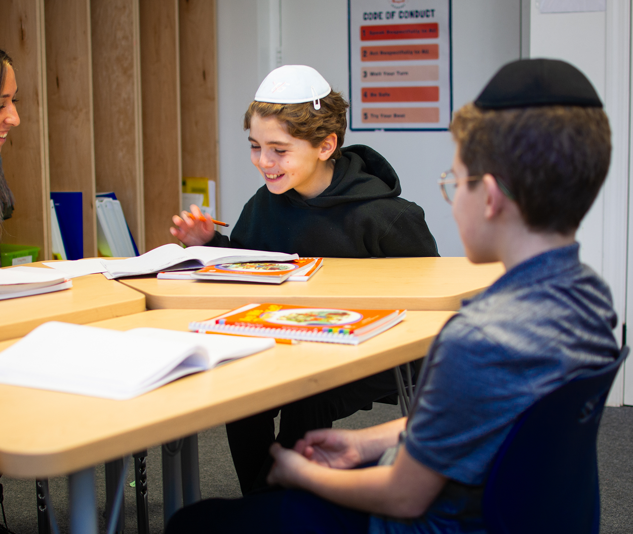 Smiling boy wearing a yarmulke studies with an open book at a classroom table with a teacher nearby.