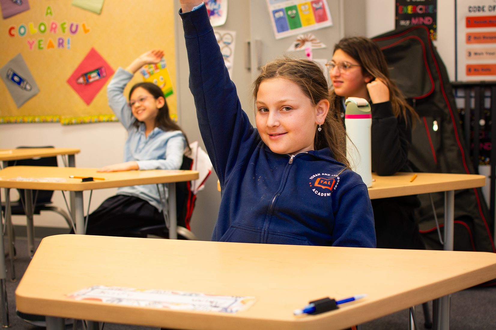 Three students at desks in a classroom, two raising their hands, one smiling at the camera.