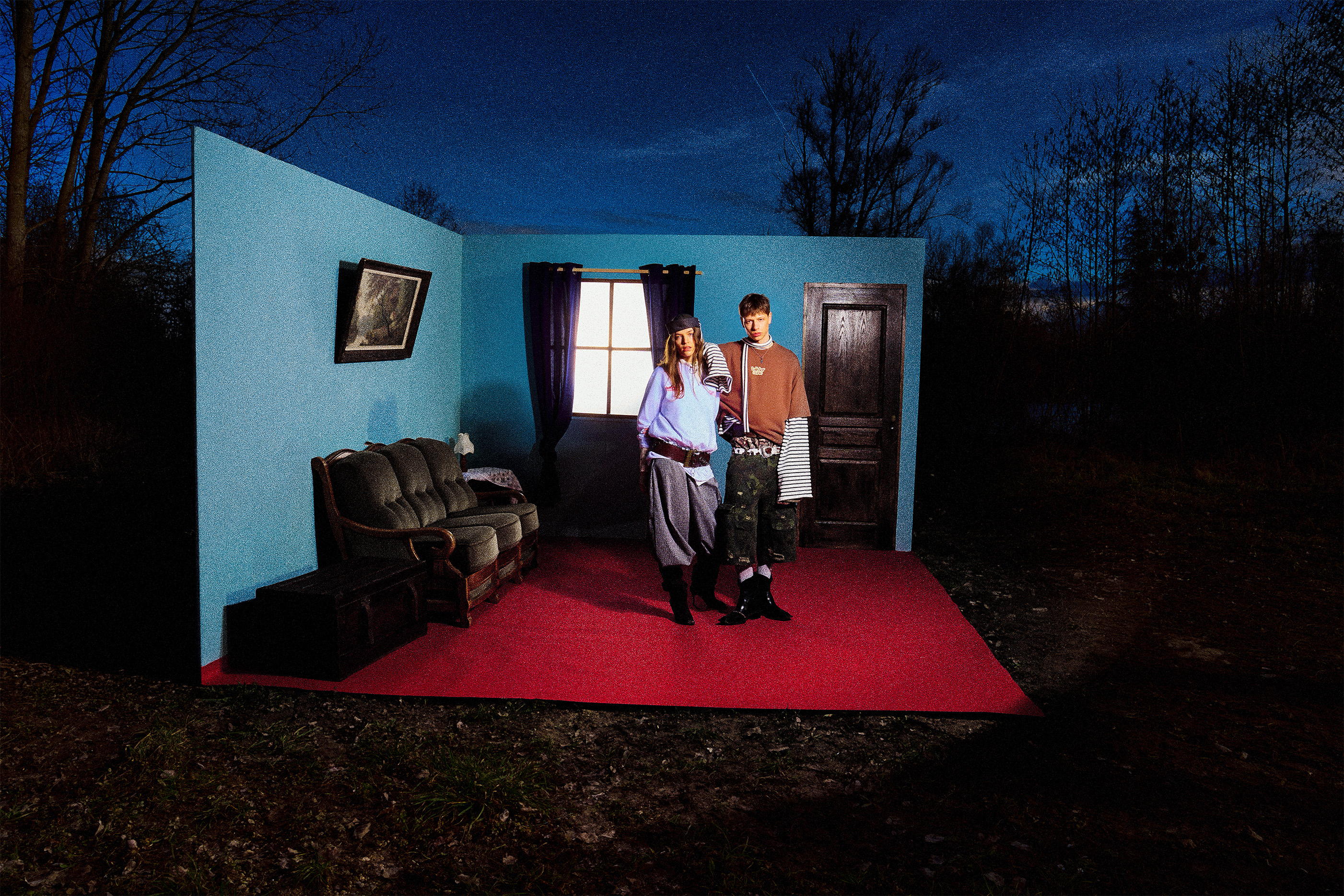 Two young people stand in a small set resembling a room with blue walls, a window with dark curtains, a door, a green couch, and a picture on the wall, all placed outdoors at night with trees in the background.