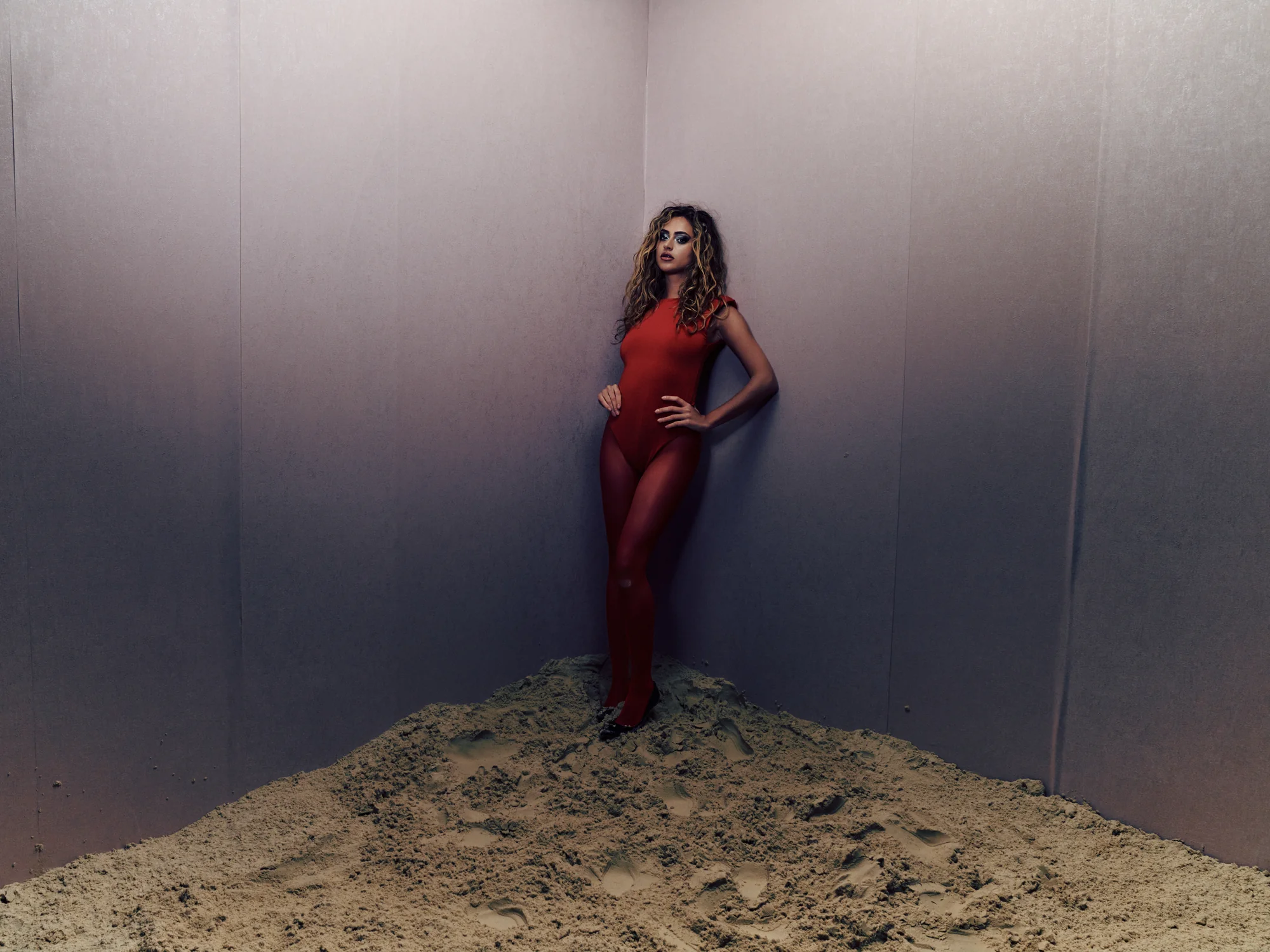 A woman with curly hair wearing a red form-fitting outfit stands confidently with one hand on her hip and the other near her waist, leaning against the corner of gray walls on a mound of sand with visible footprints.