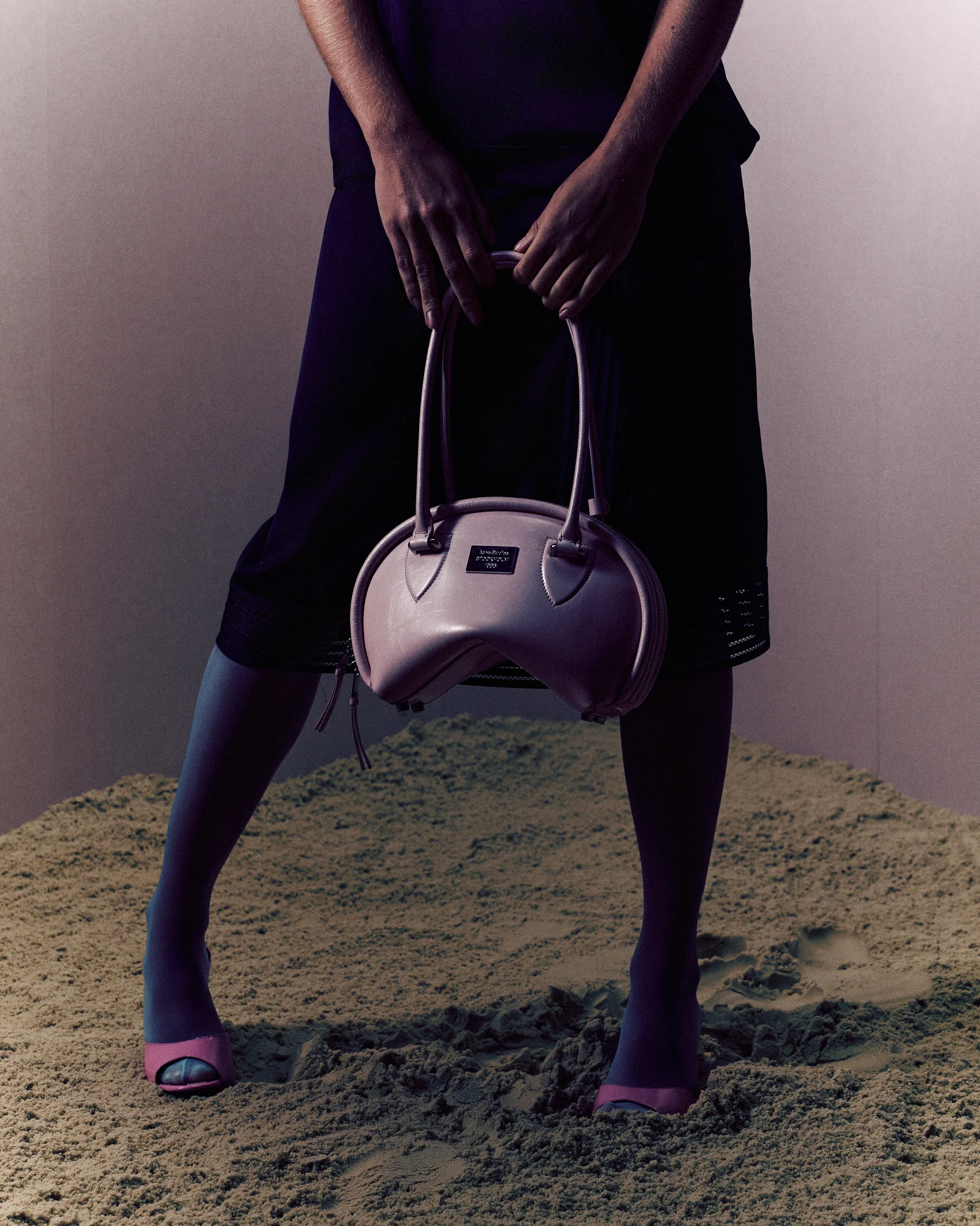 Person standing on sand wearing dark tights and pink open-toe shoes holding a uniquely shaped pink handbag with curved top handles.