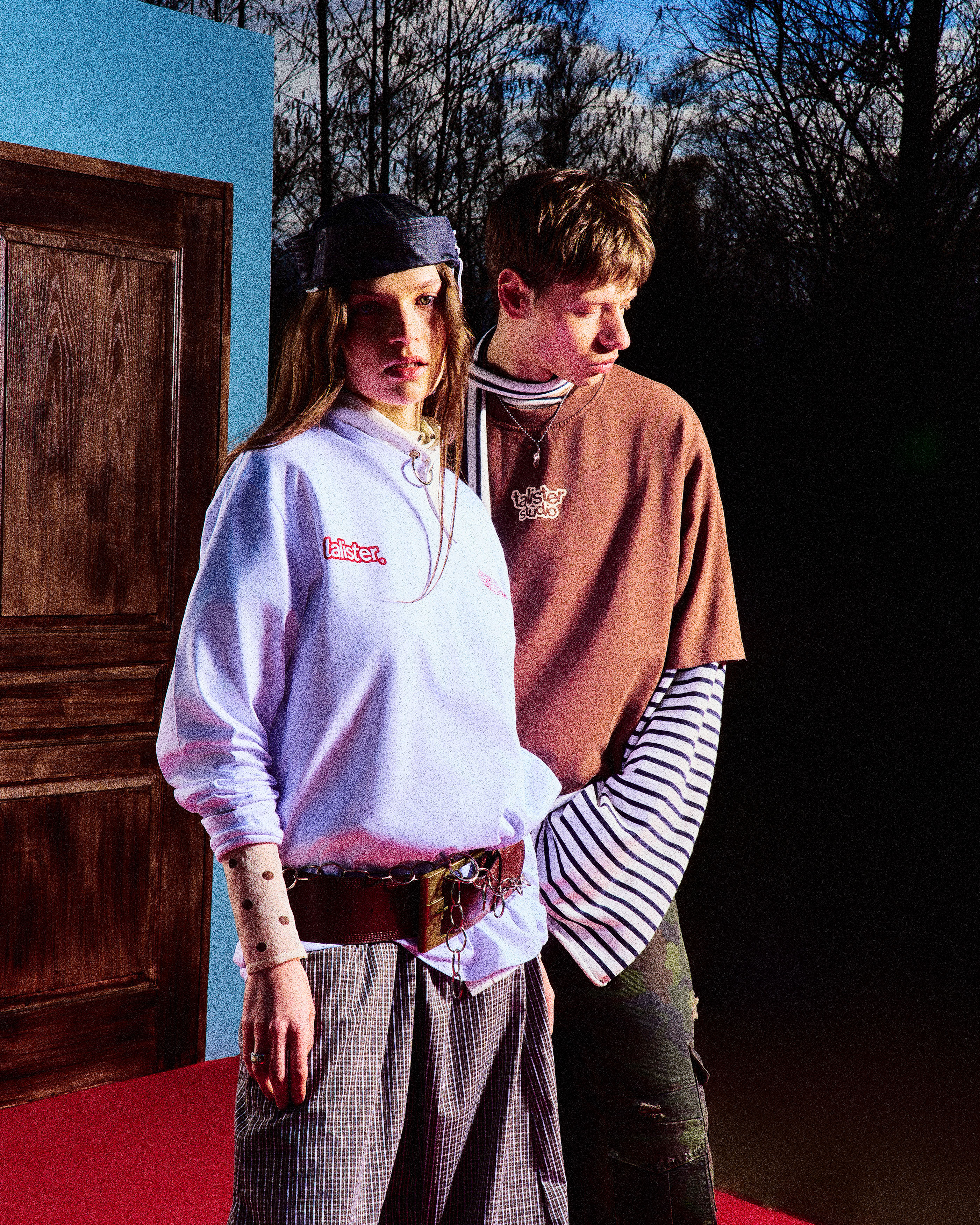 Two young people standing indoors on a red floor near a wooden door and a blue wall, wearing casual layered streetwear with graphic text on their tops. The person on the left wears a light blue sweatshirt with text, a black headband, checked pants, and a large belt with chains. The person on the right wears a brown t-shirt over a black-and-white striped long sleeve shirt and camo pants. Bare trees and evening sky are visible through a window or opening behind them.