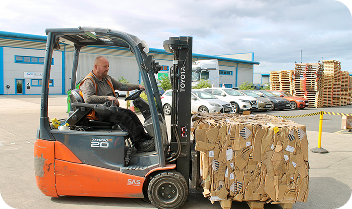 Worker operating forklift loading a large bundle of flattened cardboard boxes outdoors near cars and pallets.