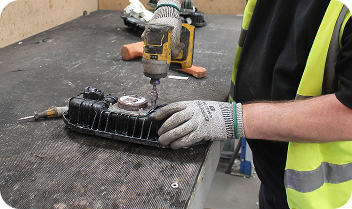 Worker in yellow vest and gloves using drill on black mechanical part on workbench.