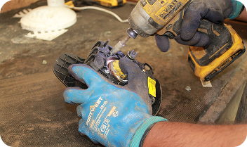 Person wearing blue gloves using a drill to repair a black plastic part on a workbench.