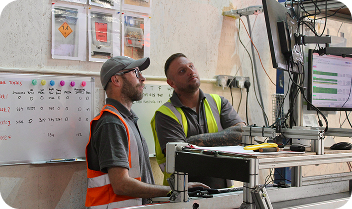 Two men wearing safety vests looking at multiple monitors in a control room.