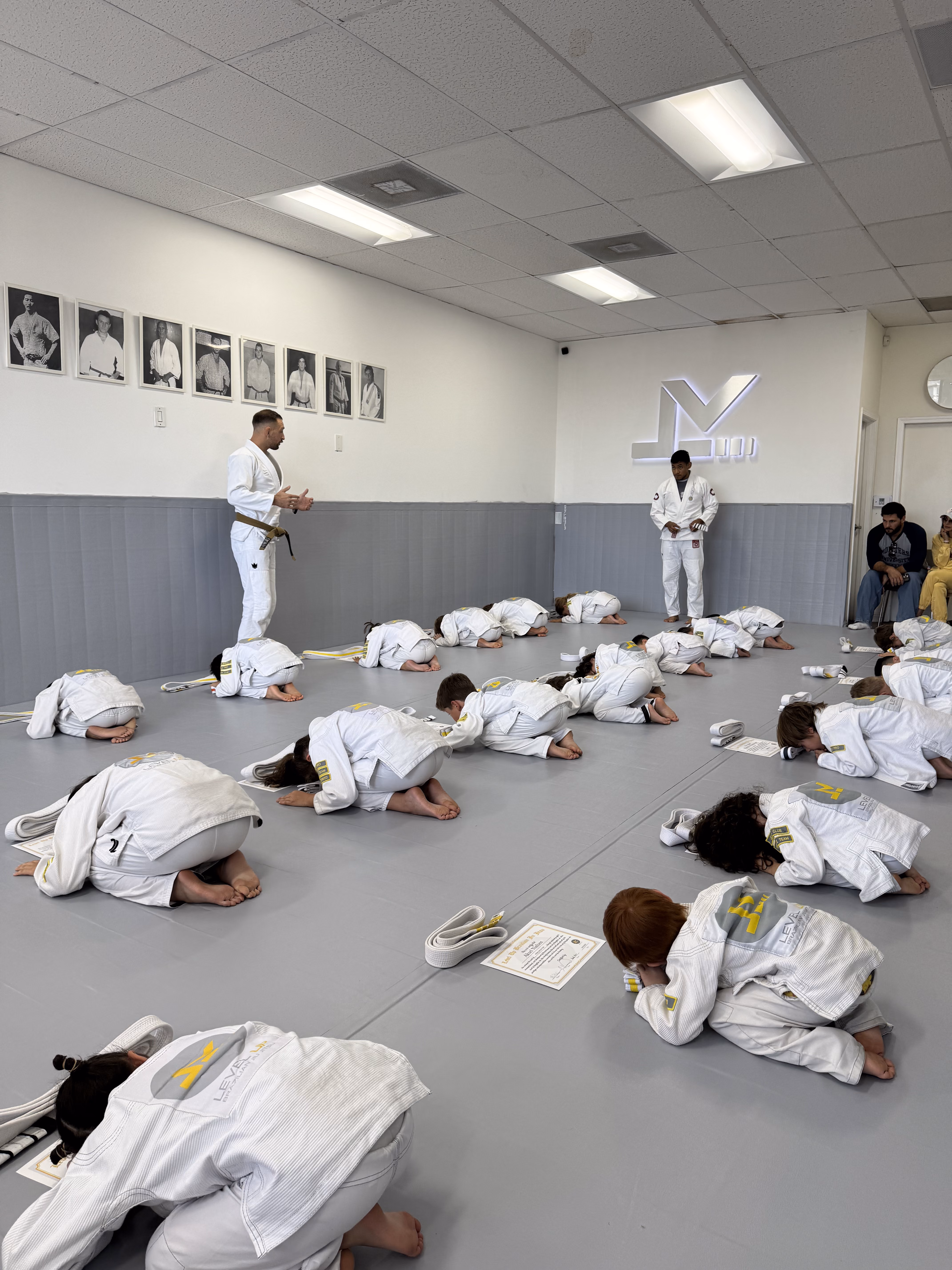 Children in white martial arts uniforms bowing on mats as two instructors stand in the front of a dojo.