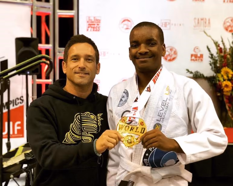 Two men smiling, one in a black hoodie, the other in a white martial arts gi holding a gold World medal.