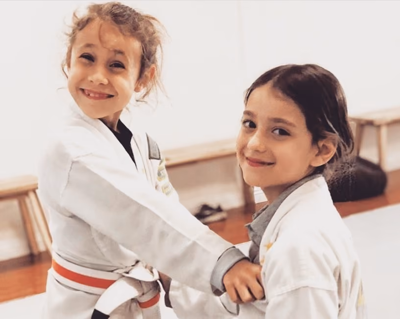 Two young girls smiling while practicing martial arts in white uniforms with belts.