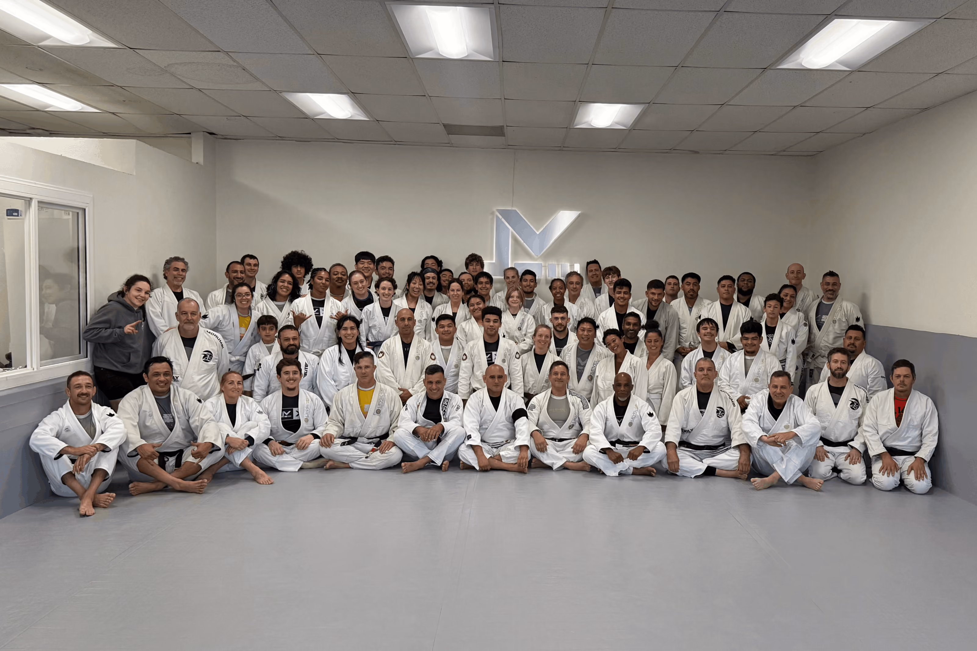 Large group of adults and teens in white martial arts uniforms posing in a dojo with a logo on the wall.