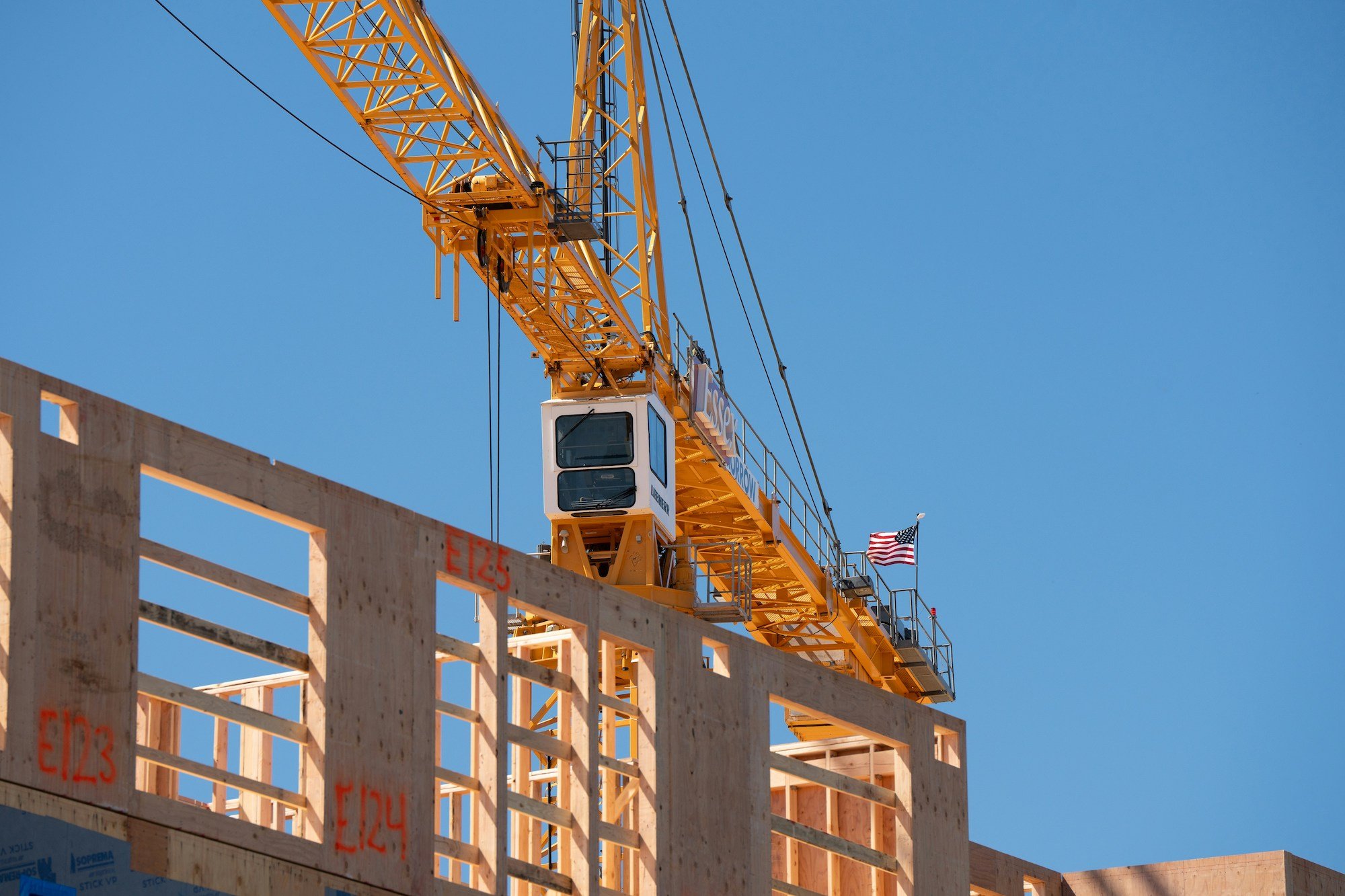 Yellow construction crane with American flag above wooden building framework under clear blue sky.